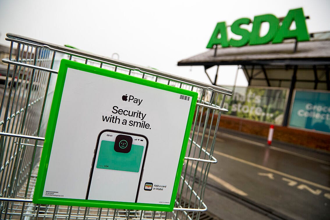 Close-up of an ASDA shopping cart featuring an Apple Pay advertisement. The ad reads “Security with a smile” with an image of an iPhone displaying Face ID and a payment card. The ASDA store signage is visible in the background.