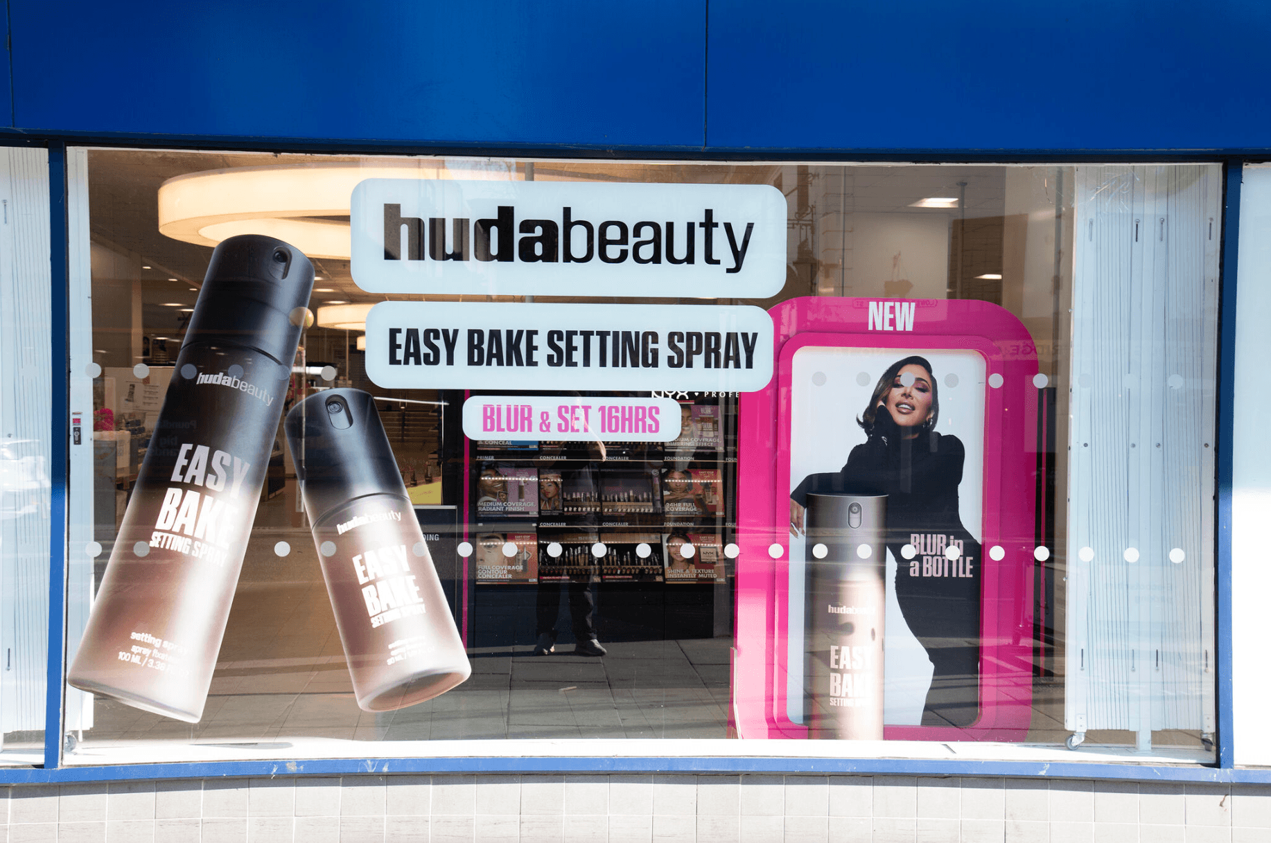 Storefront window display featuring Huda Beauty Easy Bake Setting Spray. Large bottle graphics and text highlight features such as “Blur & Set 16hrs” and “Blur in a Bottle.” A promotional image of a model appears alongside the product branding.